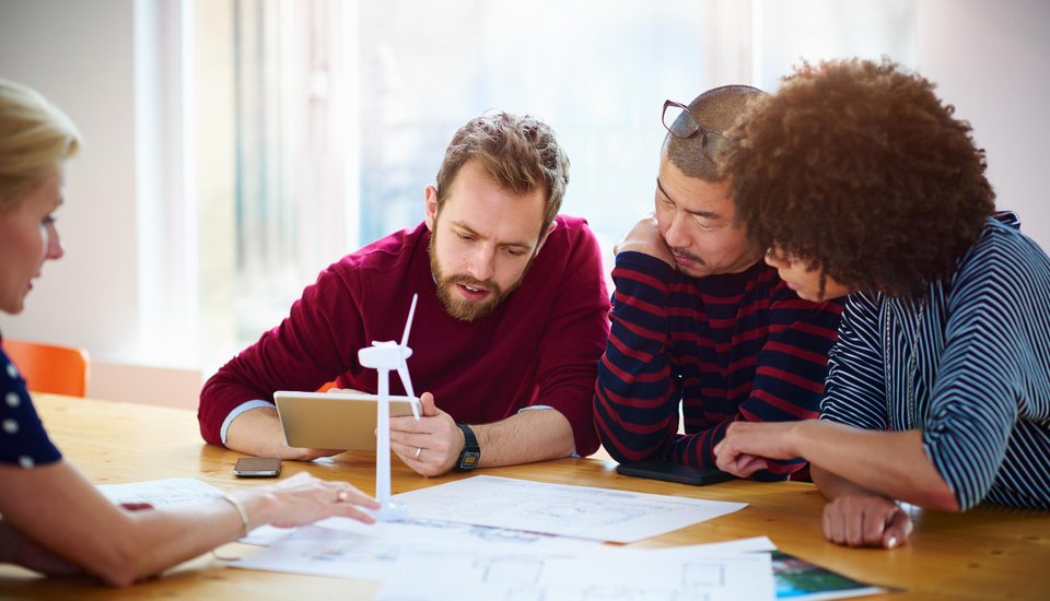 Four people sit at a table discussing documents and blueprints, with a small wind turbine model on the table. One person holds a tablet as they focus on a collaborative project in a bright room.