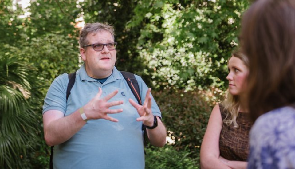 A man wearing glasses and a blue shirt gestures with his hands while talking to two women outdoors, surrounded by greenery—an engaging moment reminiscent of an RSA case study.
