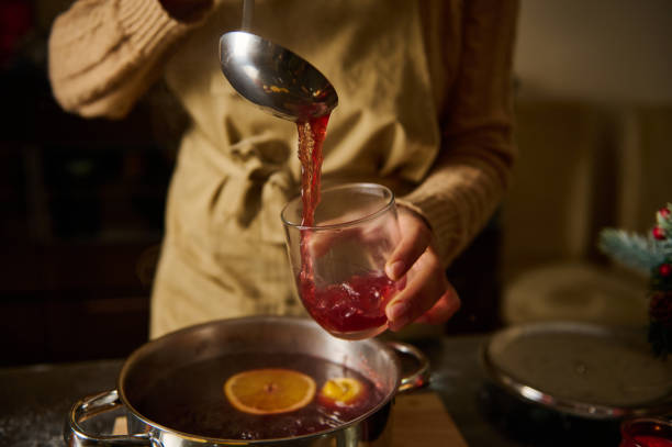 A person ladles hot mulled wine with orange slices from a pot into a glass. Wearing a beige apron and sweater, they create a warm, cozy kitchen—perfect for gathering, much like the spirit of dinings masaki sugisaki at the 2025 World Expo.