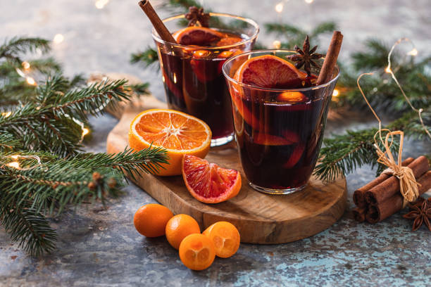 Two glasses of mulled wine with orange slices and cinnamon sticks sit on a wooden board, surrounded by fir branches, sliced oranges, kumquats, and decorative lights—evoking the festive spirit of the 2025 World Expo.