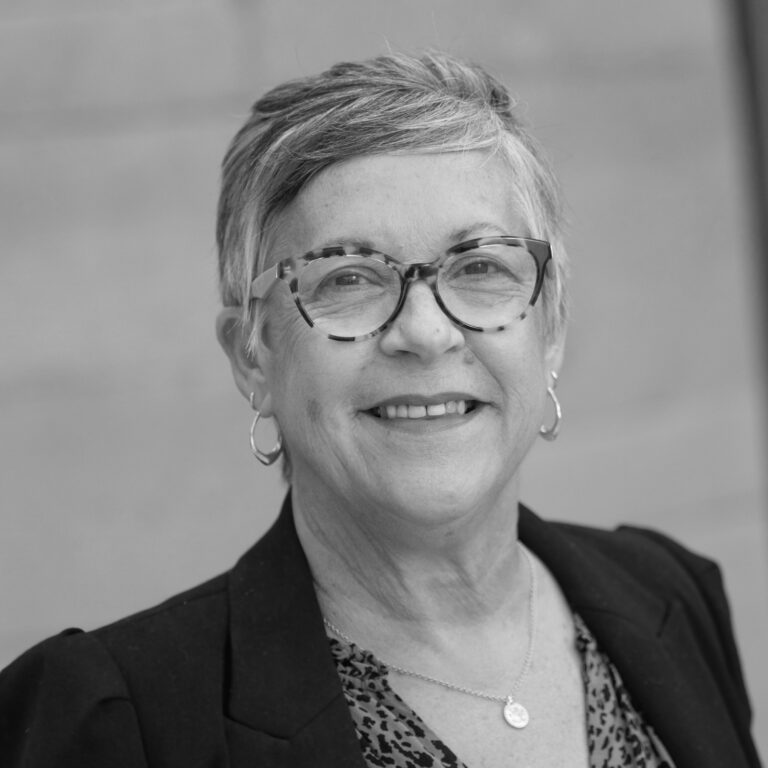 Black and white portrait of an older woman with short hair, glasses, and hoop earrings, smiling in a blazer over a patterned top—capturing the confidence found among freelancers in the creative industries, with a softly blurred background.