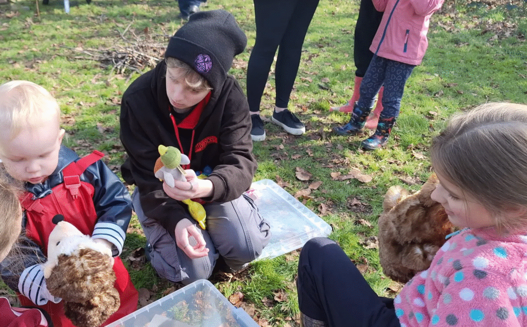 A group of children and an adult, inspired by the rsa case study approach, sit on grass outdoors holding stuffed animals and toys, with plastic boxes nearby. Some children stand in the background, and fallen leaves cover the ground.
