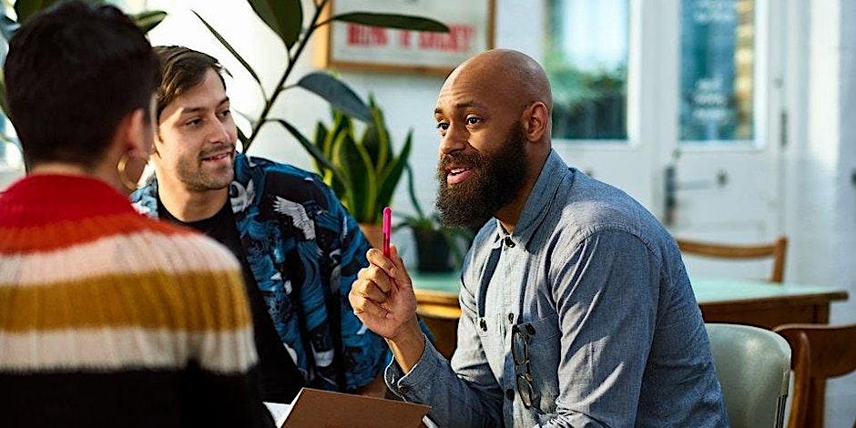 Three people are sitting together at a table in a bright room, engaged in conversation. One person is holding a pen and speaking, while the other two listen attentively. There are plants and a large window in the background.