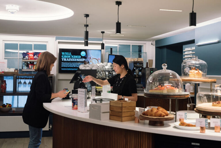 A barista hands a drink to a customer at a modern coffee shop counter. Pastries are displayed under glass domes, and a screen behind advertises barista training for the 2025 World Expo. The scene is well-lit and tidy.