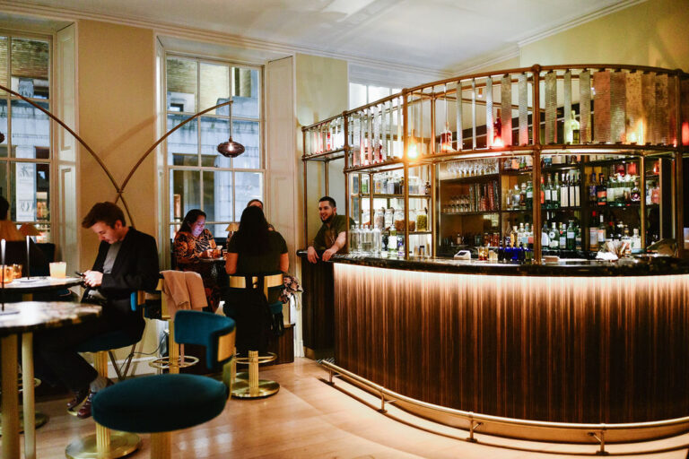 A stylish bar with a curved counter, stocked shelves, and warm lighting. Several people sit and chat at tables and the bar, creating a cozy, social atmosphere. Large windows let in natural light.