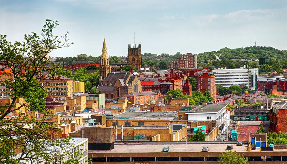 A cityscape featuring a mix of modern and historic buildings, including two prominent church towers, with lush green trees and hills in the background under a cloudy sky—perfect for discovering upcoming Birmingham events.