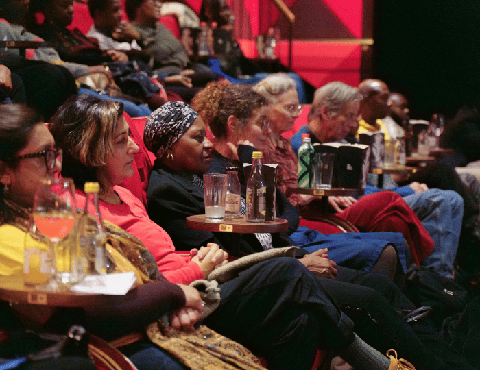 A diverse group of people sits in a theater, attentively watching a performance at the New Black Film Collective film screening. Drinks rest on small tables before them, and the seats are arranged in rows, creating a focused and engaged atmosphere.