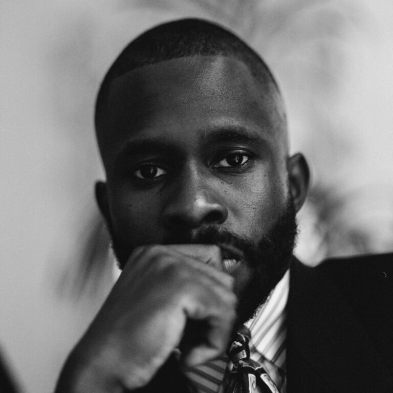 Black and white portrait of a man in a suit and tie, gazing intently at the camera with his hand resting thoughtfully near his mouth. Captured at the new black film collective film screening, the background is softly blurred.