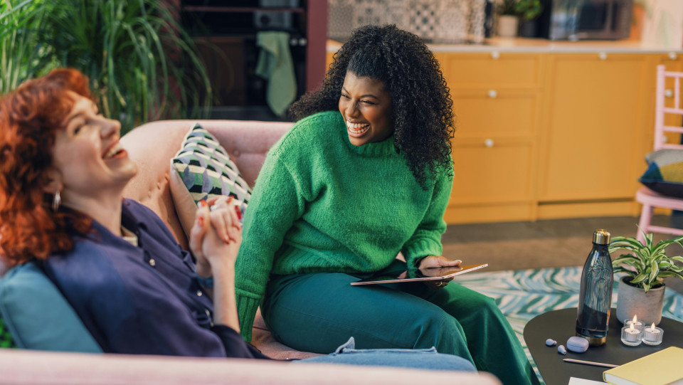 Two women sit on a sofa in a cozy living room, smiling and laughing together. Sharing stories on a tablet, they enjoy lighthearted conversation and the warmth of community. A coffee table with a plant and water bottle is nearby.
