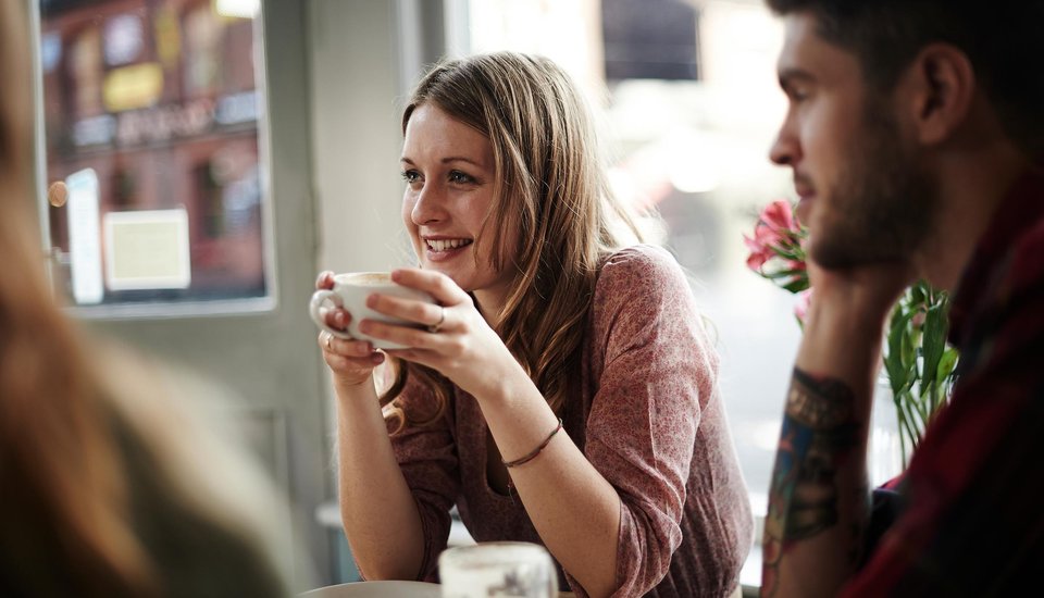 A young woman smiles while holding a cup at a café table during a Kent event, sitting across from a man with a tattooed arm. Soft natural light streams through the window, creating a warm, relaxed atmosphere.