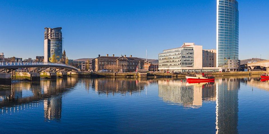 A panoramic view of a city waterfront with modern and historic buildings, a bridge on the left, and a glass skyscraper on the right—perfect scenery to explore Manchester events as red boats reflect in the calm blue water under a clear sky.