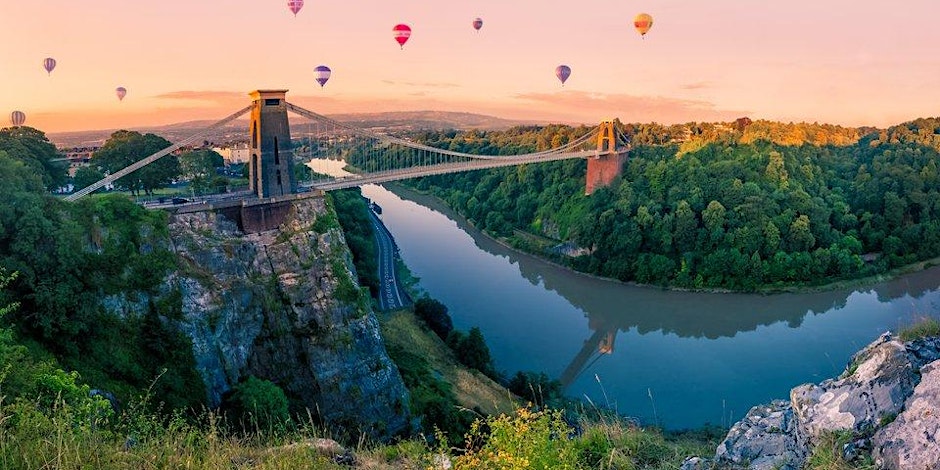 A suspension bridge spans a river between green hills at sunset, with colorful hot air balloons floating in the sky above.
