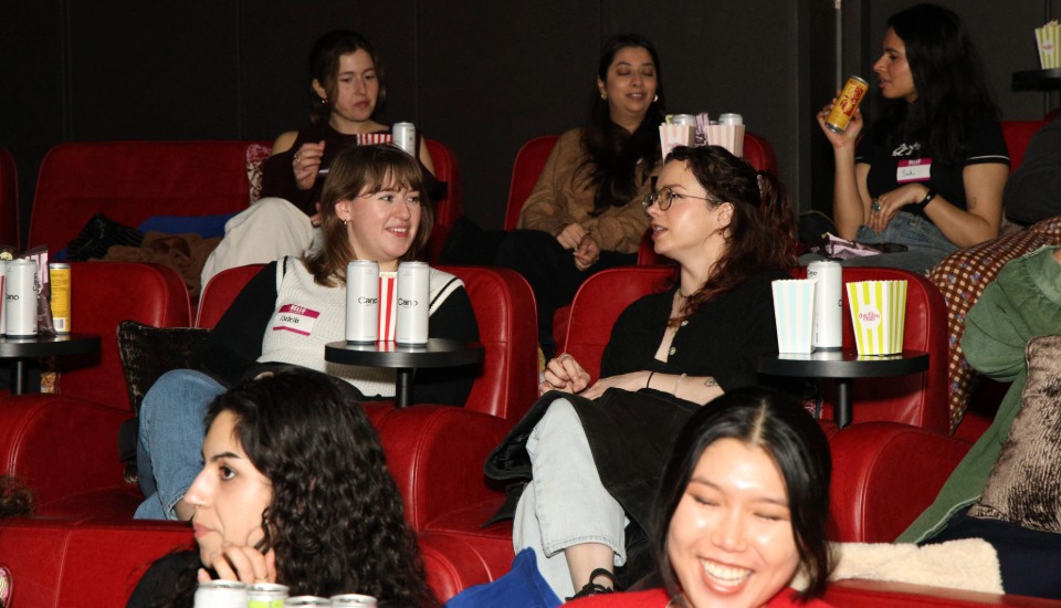 A group of women sit in red theater seats, chatting and smiling with drinks and popcorn on small tables, creating a lively and social atmosphere.