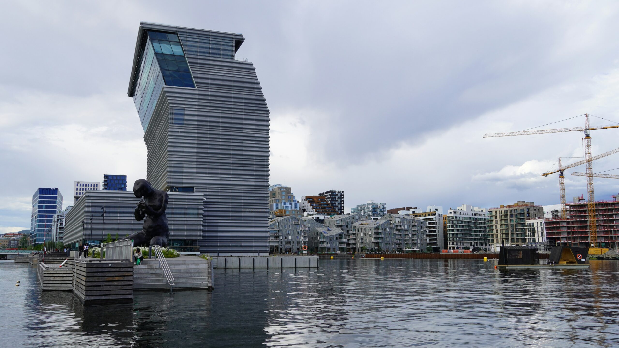 Modern waterfront cityscape with a distinctive, angular building, a large black sculpture by the water, and various contemporary apartments. Construction cranes are visible in the background under a cloudy sky.