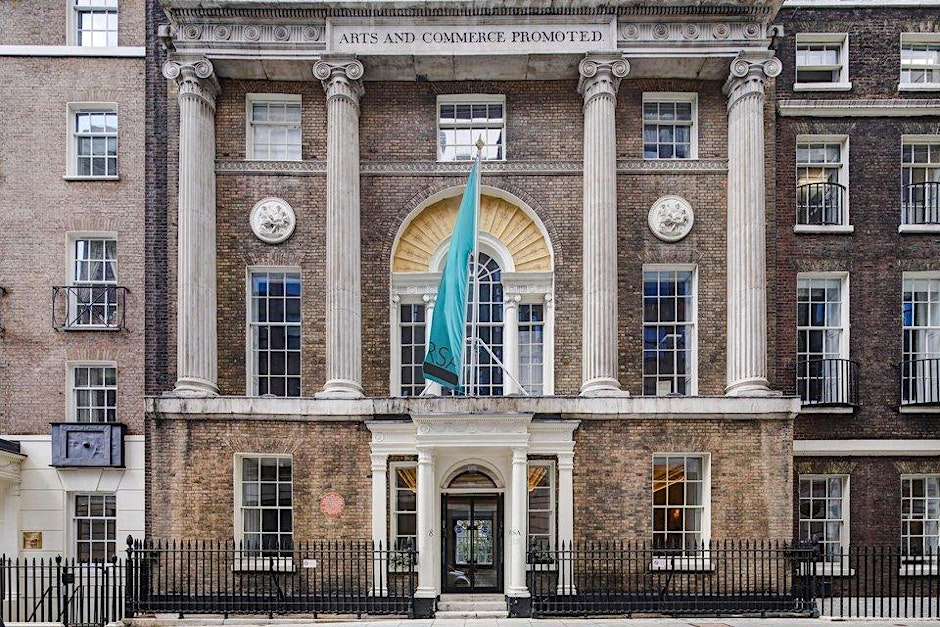 A grand, historic brick building with tall columns, large windows, and a teal flag in front. The facade features the inscription “Arts and Commerce Promoted” above the entrance.