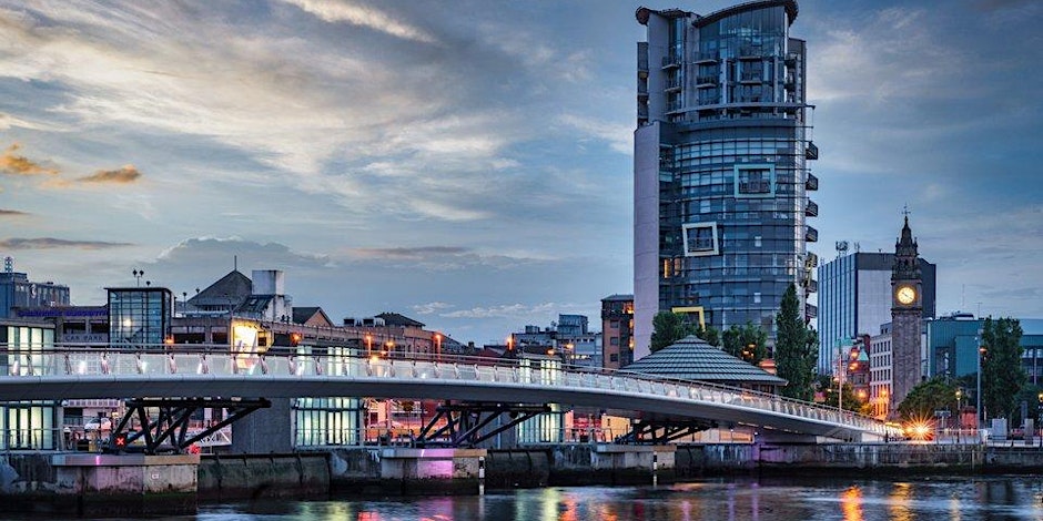 A modern pedestrian bridge crosses a river in a city at dusk, with a tall glass building and historic clock tower visible in the background under a cloudy evening sky.
