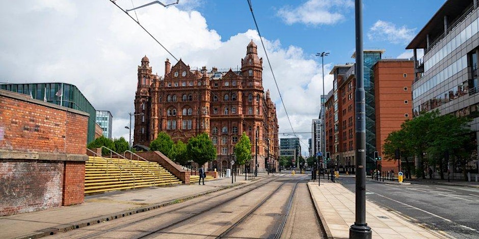 A wide street with tram tracks runs between modern buildings and a large, historic red-brick building with towers—a popular spot for Liverpool events. Steps and yellow railings line the left, with trees and a partly cloudy sky above.