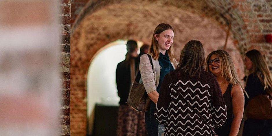 A group of women stand in a brick-arched hallway, smiling and talking with each other. Other people are visible blurred in the background. The atmosphere appears friendly and informal.