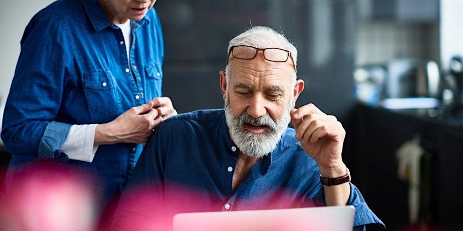 An older man with a gray beard and glasses perched on his head looks at a laptop, while a woman stands beside him. Both are wearing denim shirts, and pink flowers are blurred in the foreground.
