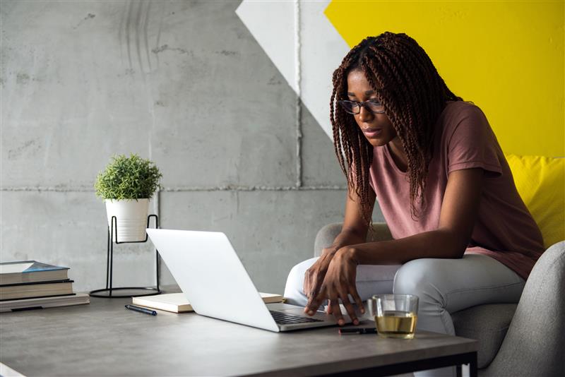 A woman with braids and glasses sits on a couch, focused on her laptop—an inspiring scene for freelancers in the creative industries. Nearby, a notebook, plant, and glass of tea rest on the table before a modern yellow and gray wall.