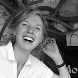 A woman with short light hair, wearing a light-colored shirt, smiles and laughs while sitting outdoors. She is holding one hand to her ear, and tree branches are visible in the background. The image is in black and white.