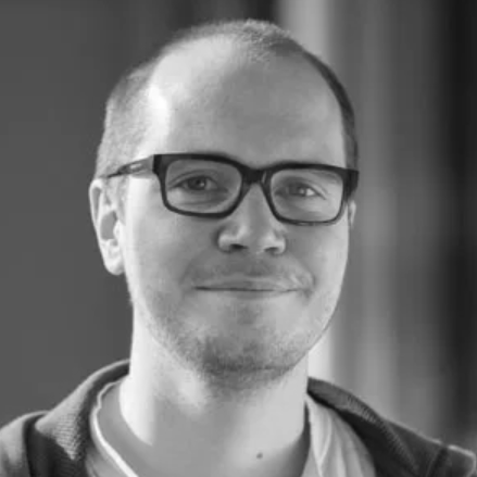 A man with short hair and glasses smiles slightly, wearing a light shirt and a dark sweater—reflecting the confident, approachable style often seen among freelancers in the creative industries. The black-and-white image features a blurred background.