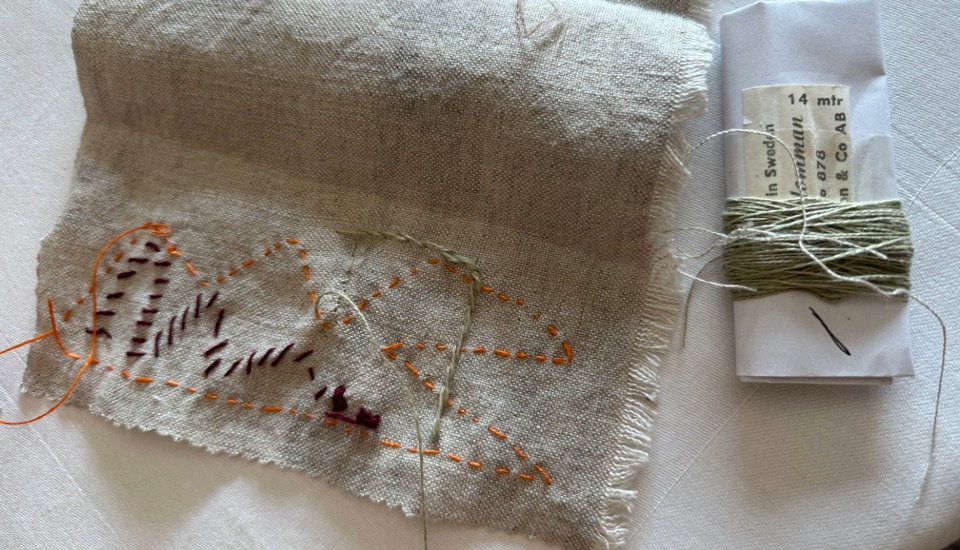 A piece of beige fabric with simple hand-stitched patterns in orange and dark thread lies next to a white card wrapped with pale green thread and a needle on a white surface.