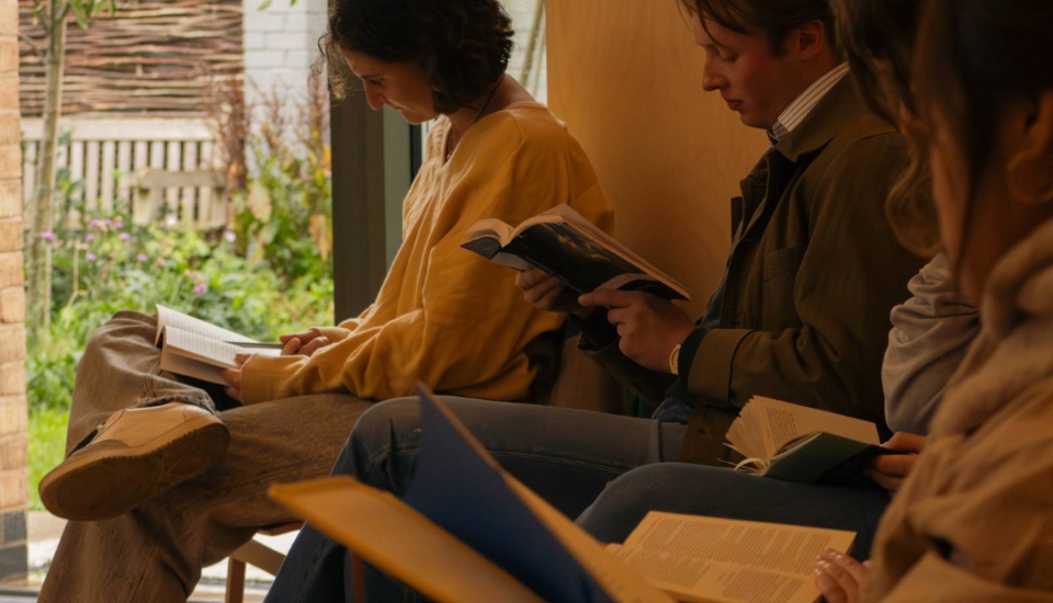 Four people sit indoors, each focused on reading a book. Sunlight filters through a nearby window, creating a warm and relaxed atmosphere. The surroundings suggest a cozy, casual setting.